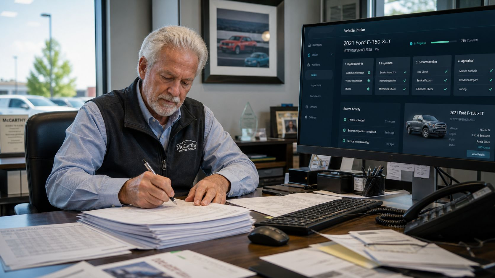 A professional man at a desk writing on documents while reviewing vehicle intake data on a computer screen.